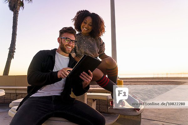 Couple sitting outdoors  beside beach  looking at digital tablet  woman wearing rollerskates