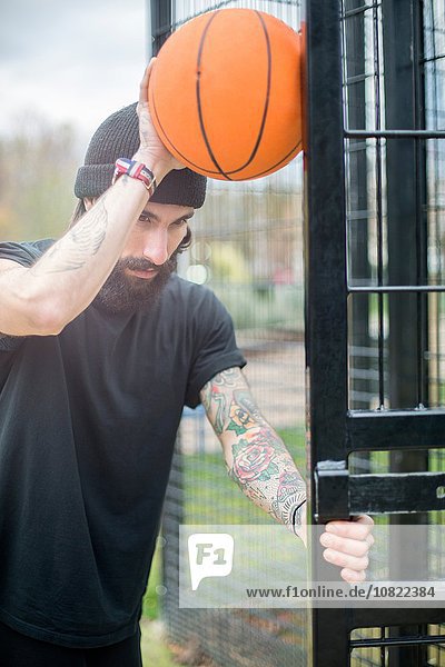 Mittlerer erwachsener Mann steht am Zaun  hält Basketball gegen Zaun  nachdenklicher Ausdruck