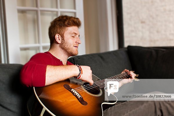 Young man sitting on sofa playing guitar  eyes closed