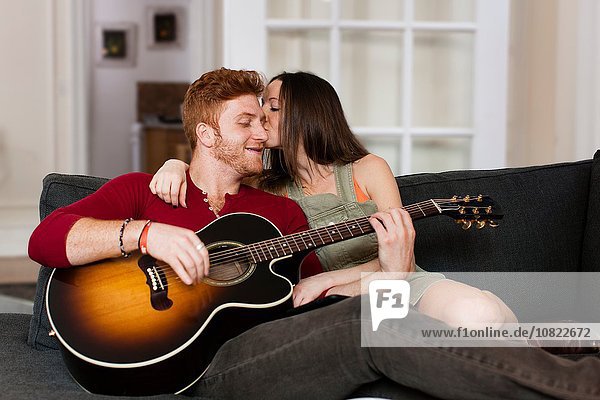 Junge Frau auf dem Sofa küsst jungen Mann  der Gitarre auf der Wange spielt.