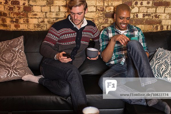 Männliches Paar sitzt auf dem Sofa und schaut auf Smartphones.