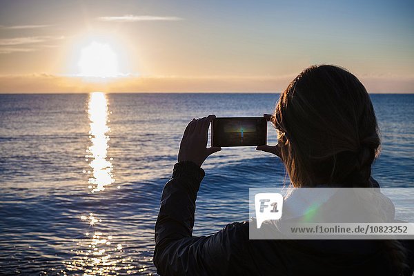Über der Schulter Silhouette einer jungen Frau  die den Sonnenuntergang über dem Meer fotografiert  Villasimius  Sardinien  Italien