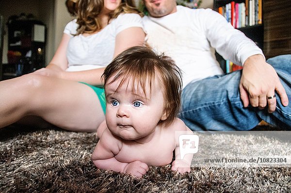 Mother and father sitting back to back on rug with baby girl lying on rug looking away