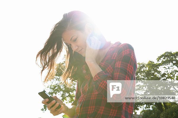 Young woman reading smartphone texts against sunlit sky