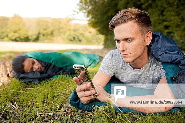 Young man lying sleeping bag texting on smartphone in field