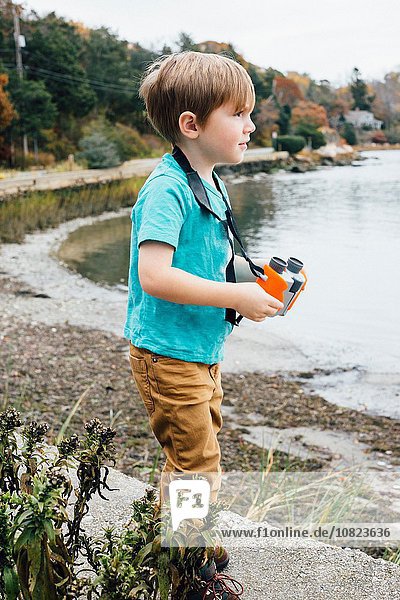 Kleiner Junge steht am See und hält ein Fernglas.