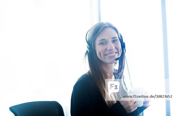 Junge Frau im Telefon-Headset mit Kaffeetasse und lächelndem Blick auf die Kamera