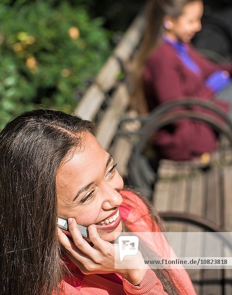 Close up of young woman sitting on park bench chatting on smartphone