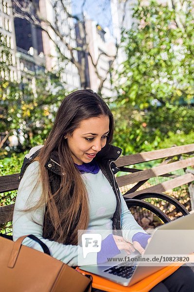 Young woman sitting on park bench typing on laptop