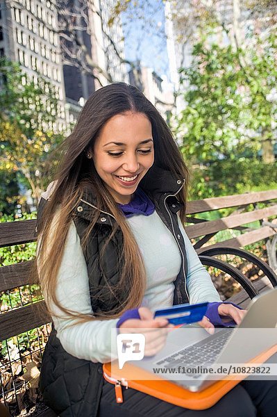 Young woman sitting on park bench using credit card with laptop