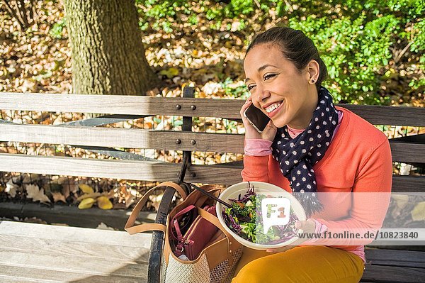 Young woman sitting on park bench chatting on smartphone whilst eating lunch