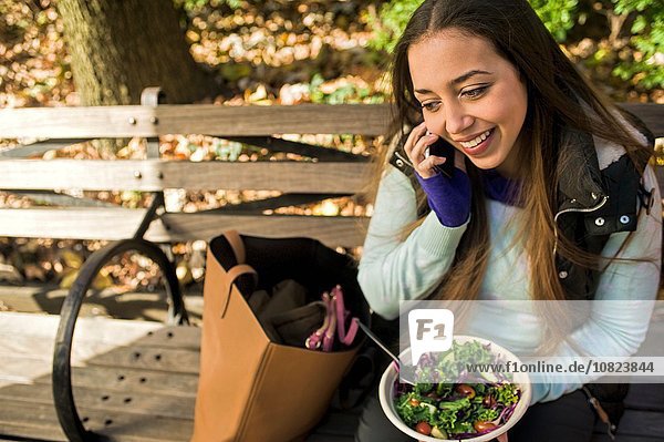 Young woman sitting on park bench chatting on smartphone whilst eating salad lunch