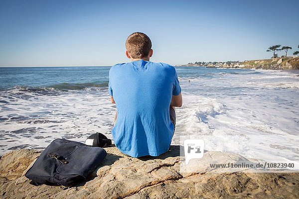 Junger Mann am Fels sitzend  Blick aufs Meer  Rückansicht