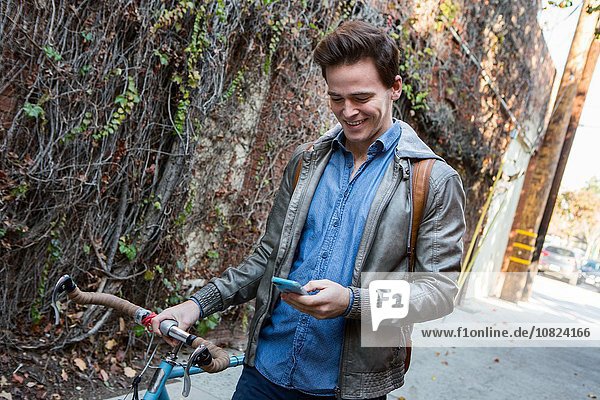 Young man strolling with cycle on sidewalk reading smartphone texts