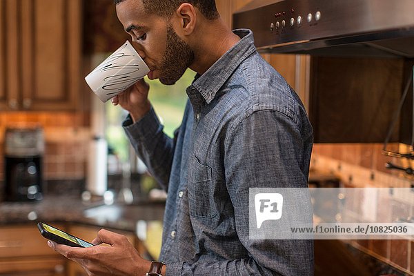 Mittlerer Erwachsener Mann in der Küche liest Smartphone-Text und trinkt Kaffee.