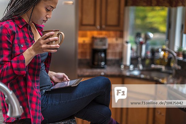 Mittlere erwachsene Frau  die in der Küche sitzt und ein digitales Tablett benutzt.