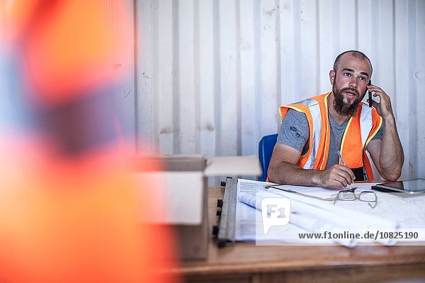 Construction worker sitting at desk talking on smartphone in portable cabin