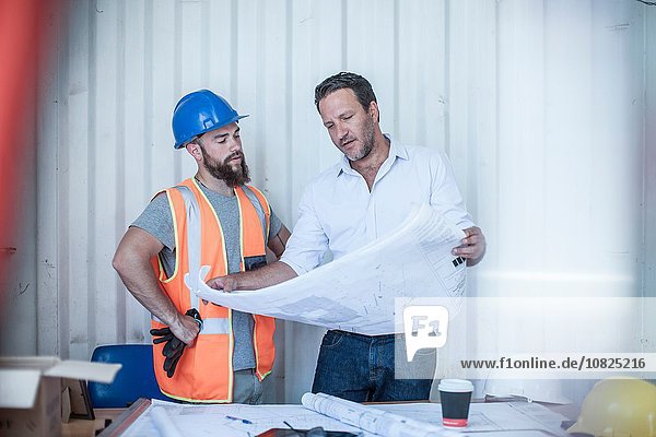Construction foreman looking over blueprint with worker at desk in portable cabin