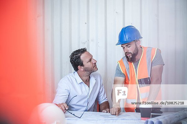Worker explaining blueprint to construction foreman at desk in portable cabin