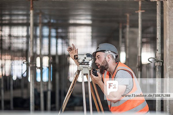 Young male surveyor looking through theodolite giving hand signal on construction site