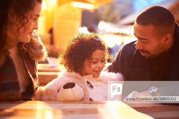Girl at amusement park holding teddy bear sitting at table with parents