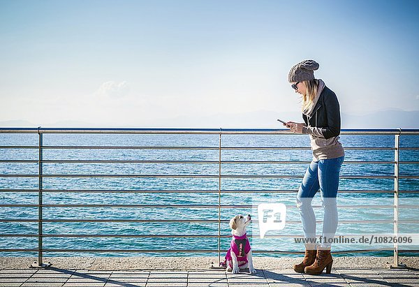 Durchgehende Seitenansicht der jungen Frau mit Hund am Geländer vor dem Meer mittels Smartphone