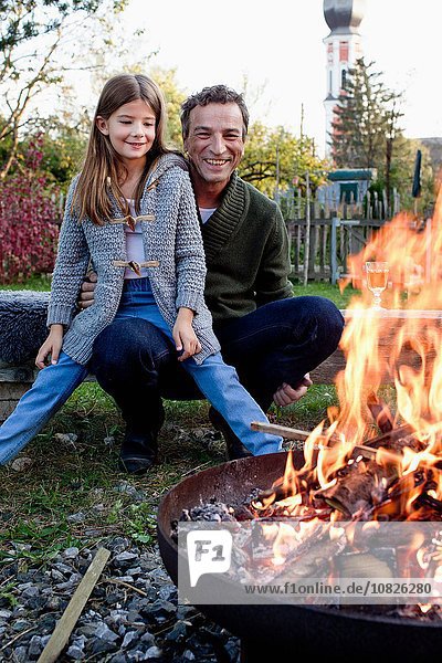 Girl sitting on father's lap in garden watching fire pit