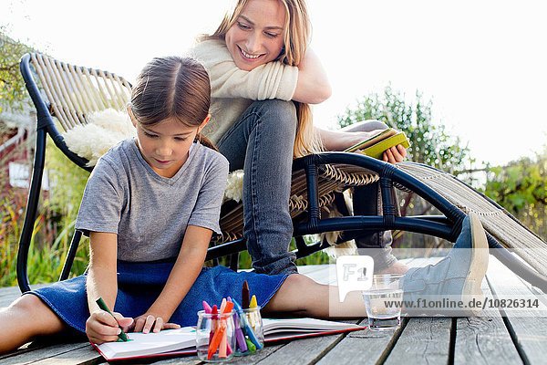 Girl drawing on decking with mother sitting close by  smiling