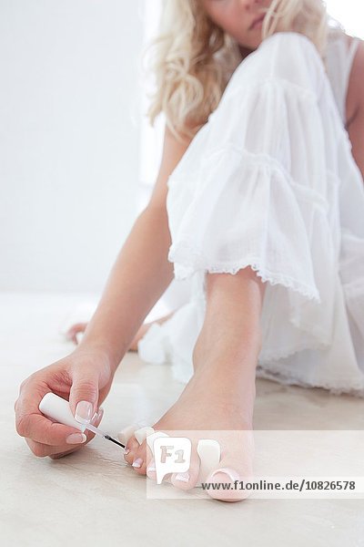 Ausschnitt einer jungen Frau  die auf dem Boden sitzt und Nagellack auf die Zehennägel aufträgt.