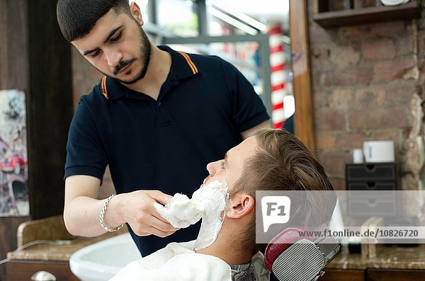Junger Mann im Friseurladen  der Rasiercreme auf das Gesicht des Kunden aufträgt.