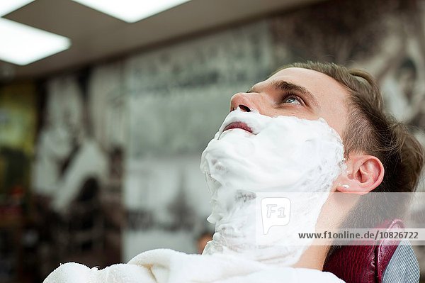Junger Mann im Friseurladen mit einem Gesicht  das mit Rasierschaum bedeckt ist.