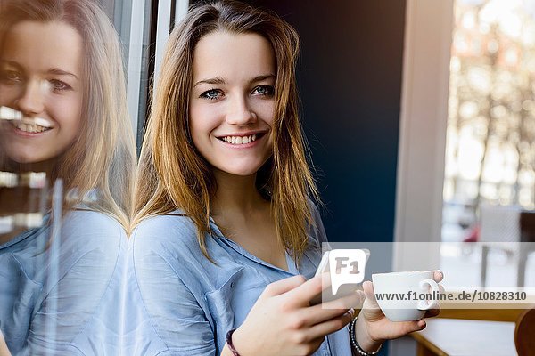 Young woman holding coffee cup and smartphone looking at camera smiling
