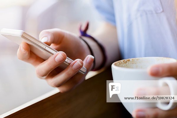 Beschnittene Ansicht von jungen Frauenhänden  die eine Kaffeetasse mit dem Smartphone halten.