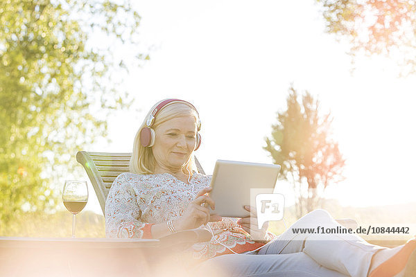 Seniorin mit Kopfhörer und Wein mit digitalem Tablett auf Sessel im sonnigen Hinterhof