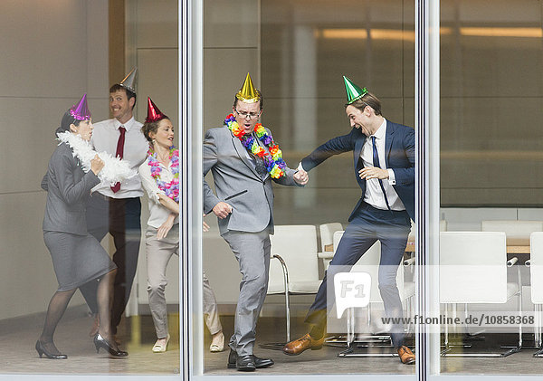 Verspielte Geschäftsleute in Partyhüten tanzen am Konferenzraumfenster