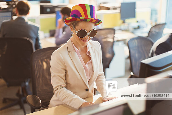 Playful businesswoman wearing silly sunglasses and striped hat at computer in office