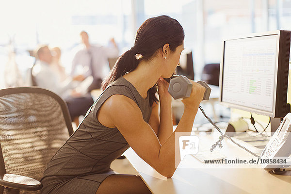 Businesswoman doing biceps curls with dumbbell and talking on telephone at computer in office