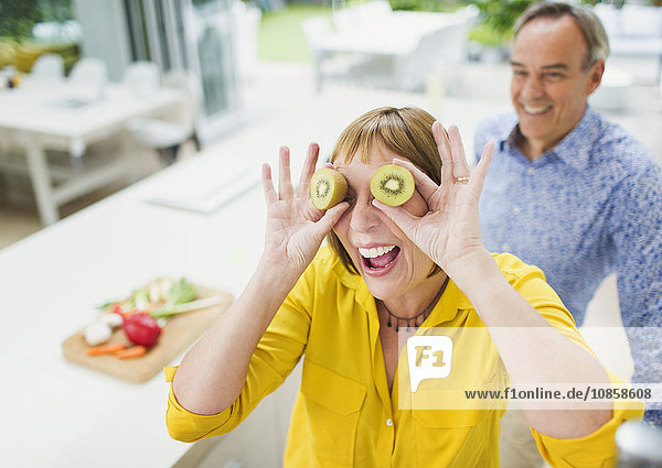 Portrait playful mature woman covering eyes with kiwi slices