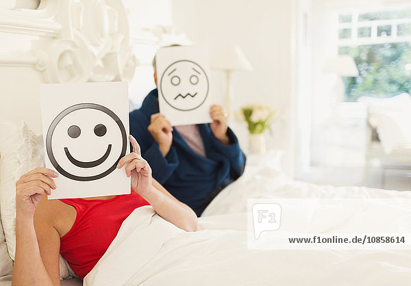 Portrait of couples holding smiling face and frowning face printouts in bed