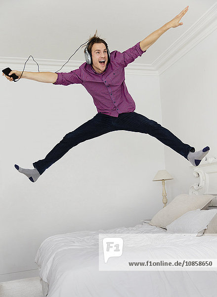 Playful man jumping on bed with legs apart listening to music with mp3 player and headphones