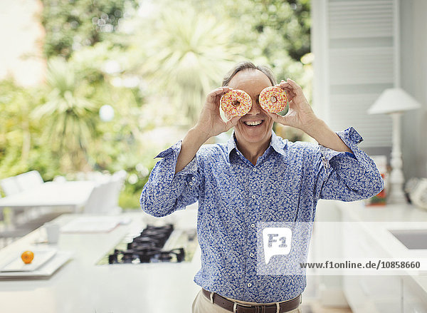 Portrait playful mature man covering eyes with donuts
