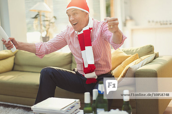 Enthusiastic mature man in hat and scarf watching TV sports event on living room sofa