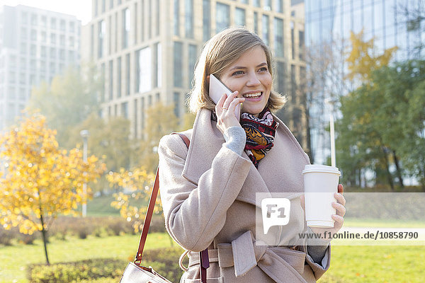 Lächelnde Geschäftsfrau mit Kaffee am Handy im Stadtpark
