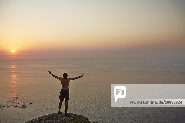 Exuberant male runner with arms outstretched on rock overlooking ocean sunset