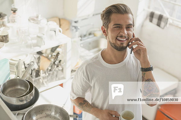 Tattooed young man talking on cell phone and drinking coffee in apartment kitchen