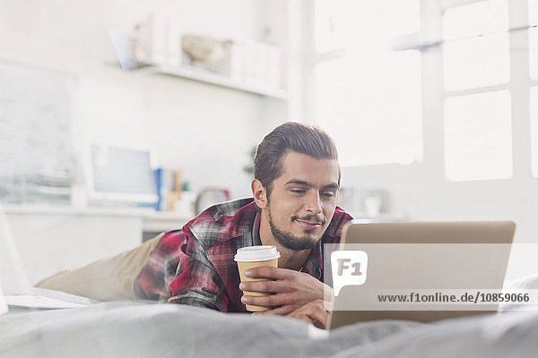 Young man drinking coffee at laptop on bed