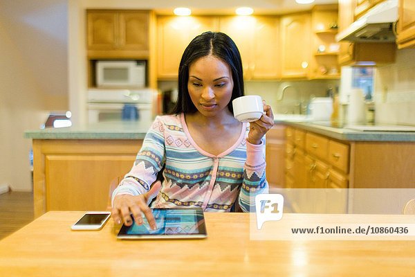 Junge Frau trinkt Kaffee und benutzt digitalen Tablet-Touchscreen am Küchentisch