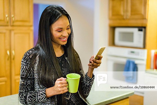 Junge Frau trinkt Kaffee in der Küche und liest dabei Text auf einem Smartphone