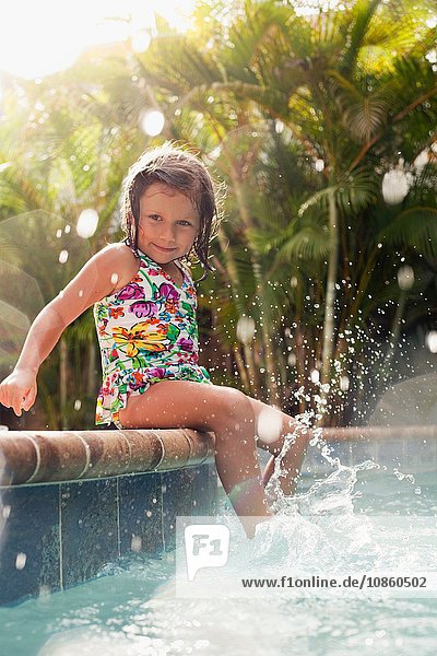 Girl wearing swimming costume sitting on sunlit poolside splashing