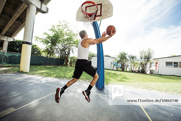 Seitenansicht eines jungen Mannes auf einem Basketballfeld in der Luft  der Basketball für einen Reifen springen lässt